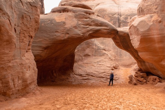 girl in blue jacket and white hat looking up at sand dune arch standing in sand in Arches national Park on a Southwest Road Trip