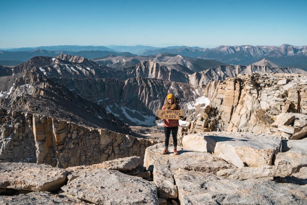 girl in pink jacket and yellow hat holding Mount Whitney elevation sign at Mount Whitney summit