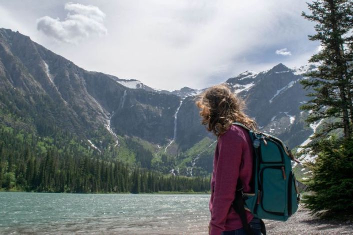 girl in pink sweater with blue backpack looking at a lake with waterfalls and her hair is blowing back from the wind