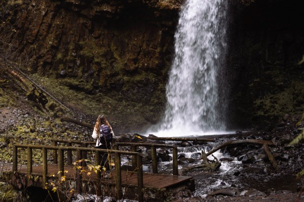 girl in white coat and purple backpack on bridge looking at Upper Latourell Falls