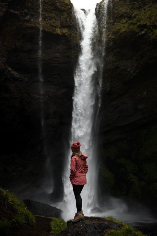 girl in red jacket and hat standing in front of waterfall that is pouring into a canyon
